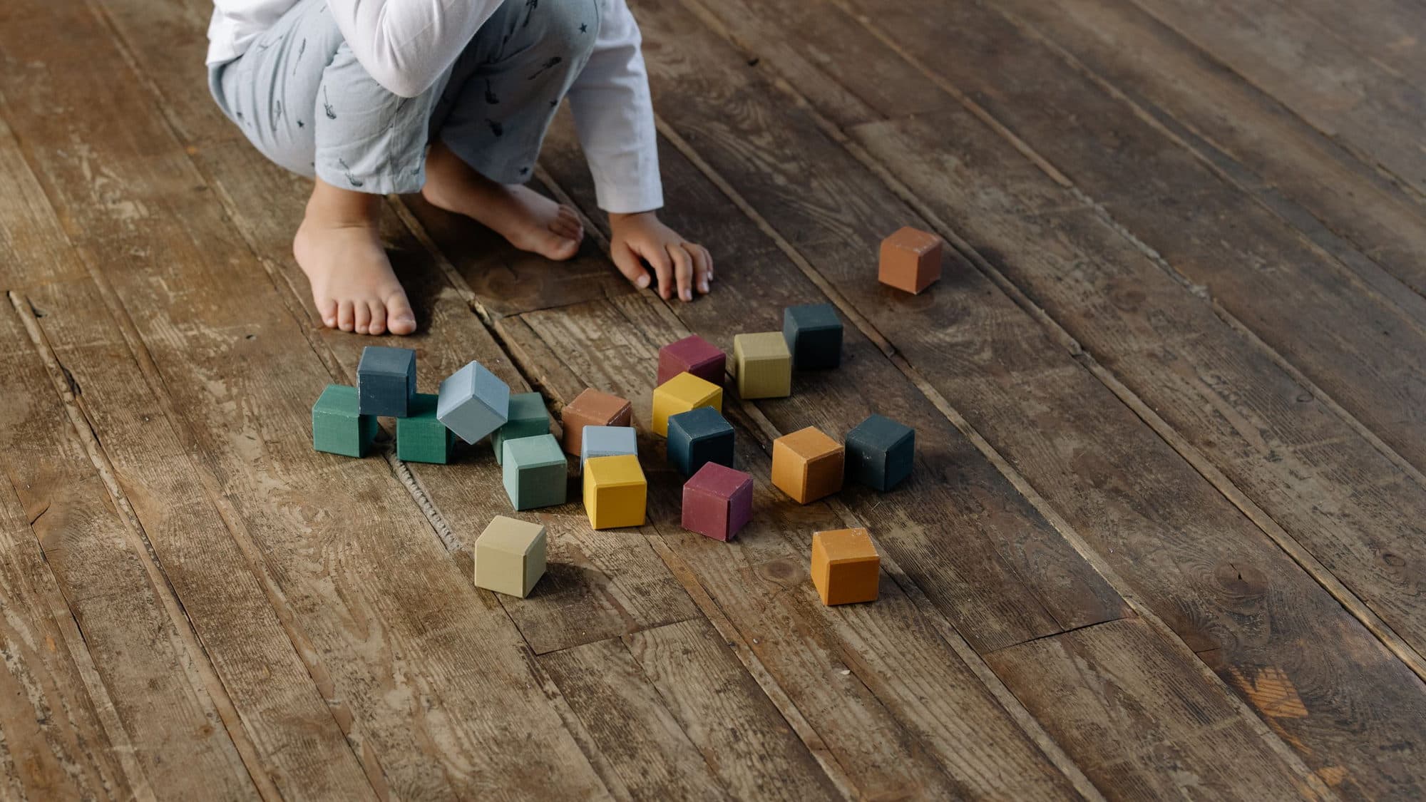 Bé ngồi xổm trên sàn gỗ mộc, bày rải quanh chân là những khối gỗ vuông màu trầm đang chơi dở