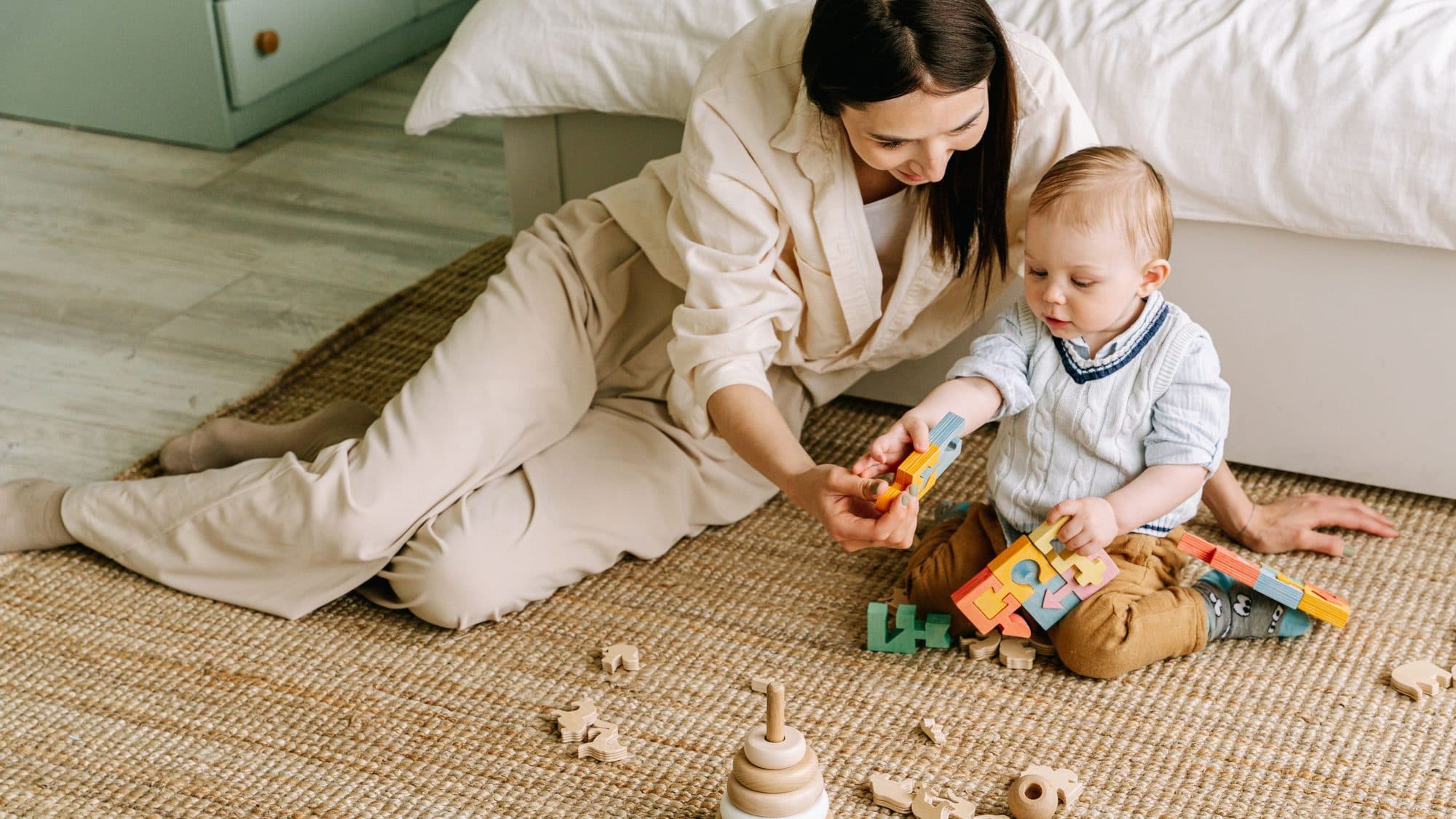 Người mẹ ngồi bệt trên thảm cói, đưa cho bé một miếng ghép gỗ; quanh hai mẹ con là những hình thú gỗ và tháp vòng gỗ đang chơi dở